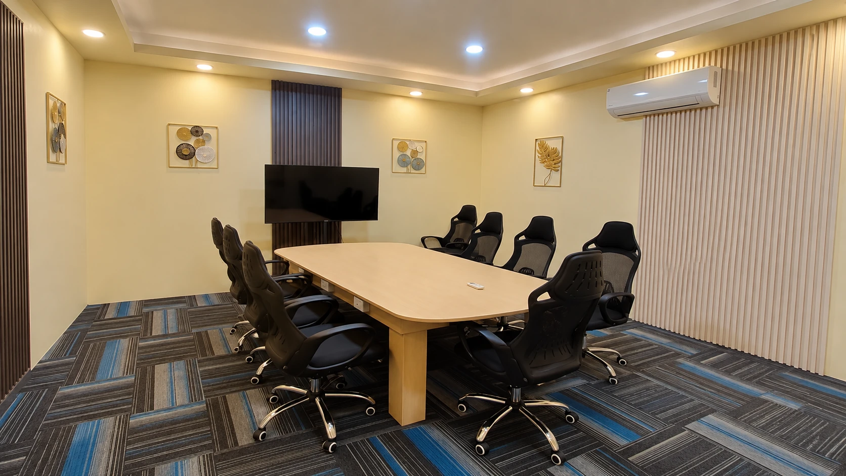 A conference boardroom with a long table, black chairs, and a wall-mounted screen.
